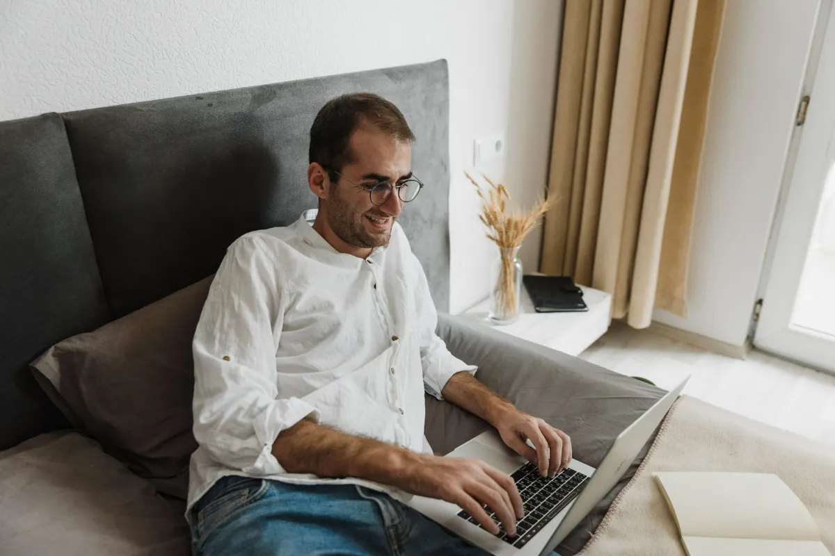 A man in glasses browsing on a laptop while sitting on a bed